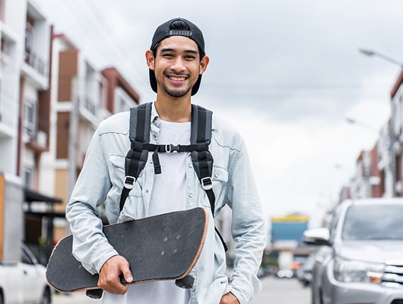 A young man wearing a backpack and smiling while holding a skateboard outdoors.